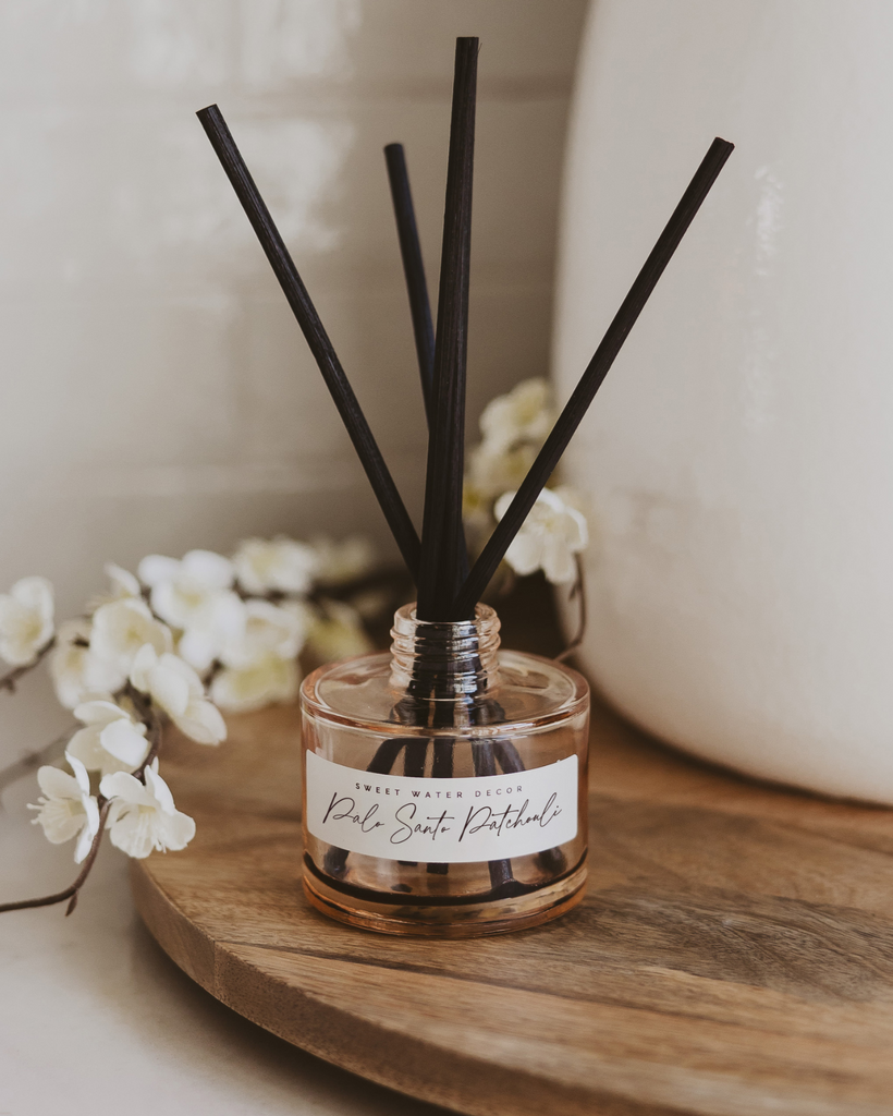 Reed diffuser with black sticks in a glass bottle on a wooden surface with white flowers in the background.