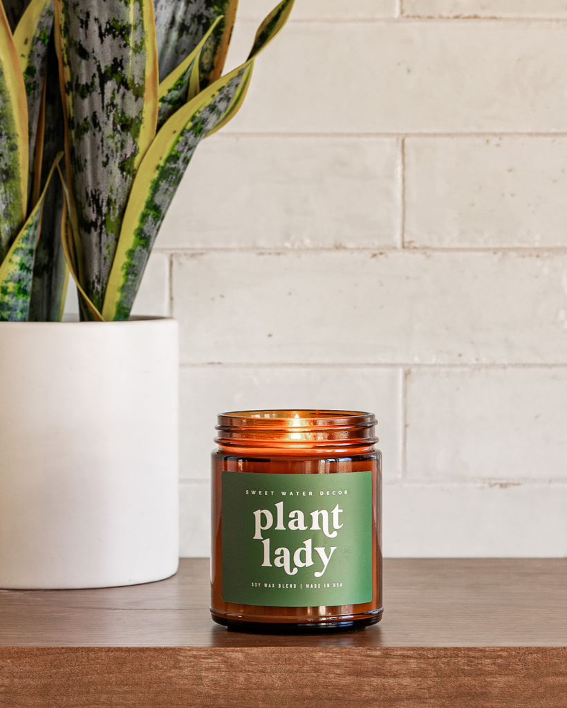 Candle labeled 'plant lady' next to a potted plant on a wooden surface with a tiled wall background.