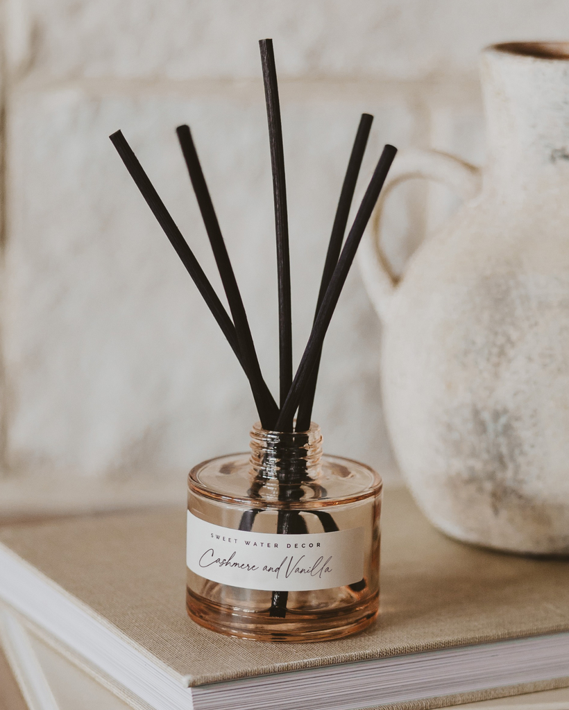 Reed diffuser with black sticks in a clear container on a book with a vase in the background